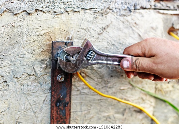 Master electrician\
connecting ground wire to the metal rod near dwelling house.\
Electrical worker hands fixing cable to earth bonding bar with\
adjustable wrench or gas\
wrench