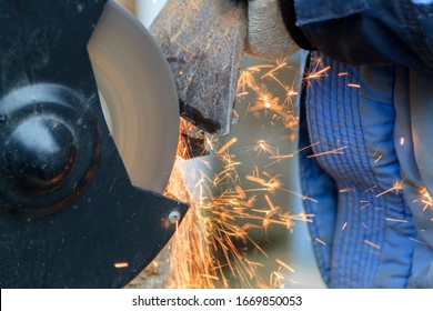 Master In Gloves And Blue Jacket Sharpens Axe On Electric Sharpener Outdoors. Process Of Sharpening Blunt Tool With Sparks Flying In Different Directions