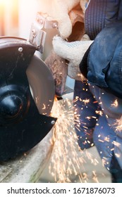 Master In Gloves And Blue Jacket Sharpens Axe On Electric Sharpener Outdoors. Process Of Sharpening Blunt Tool With Sparks Flying In Different Directions