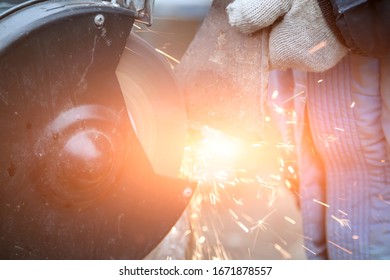 Master In Gloves And Blue Jacket Sharpens Axe On Electric Sharpener Outdoors. Process Of Sharpening Blunt Tool With Sparks Flying In Different Directions