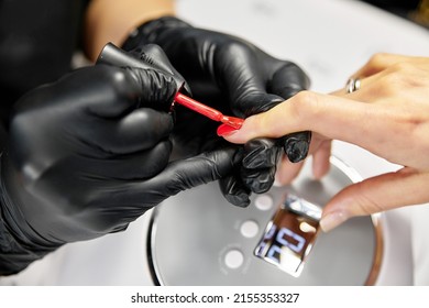 the master makes a manicure with a gel polish coating for a woman with clean nails. a master in black latex gloves paints a nail with red varnish