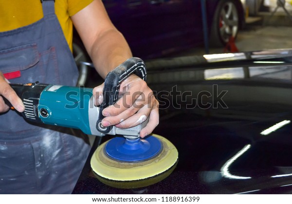 The master man of the detailing in work clothes and\
dirty hands polishes the bodywork of the bonnet of the car in black\
with a polishing machine in the workshop for body repair\
cars