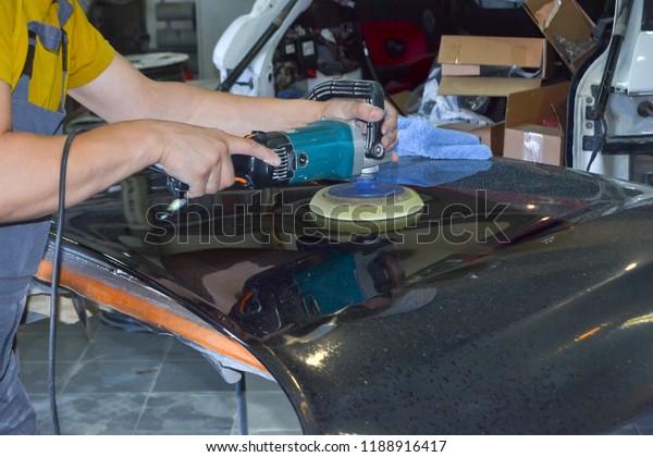 The master man of the detailing in work clothes and\
dirty hands polishes the bodywork of the bonnet of the car in black\
with a polishing machine in the workshop for body repair\
cars