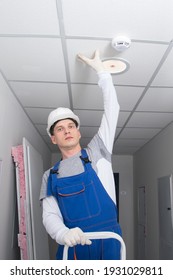 A Master From The Management Company, Wearing A Hard Hat And A Professional Uniform, Was Replacing A Light Bulb And Checking The Fire Alarm System In The Space Of The Suspended Ceiling