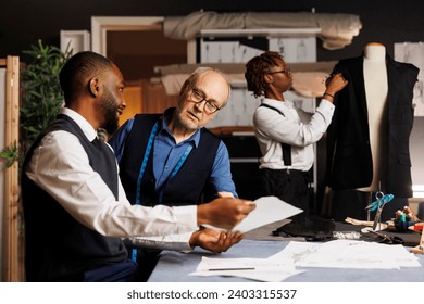 Master tailor working with diverse apprentice team of suitmakers to finalize client sartorial comission, checking outfit sketches in professional atelier. Couturiers manufacturing bespoken clothing