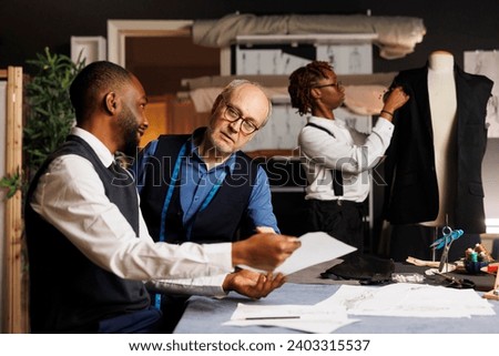 Master tailor working with diverse apprentice team of suitmakers to finalize client sartorial comission, checking outfit sketches in professional atelier. Couturiers manufacturing bespoken clothing