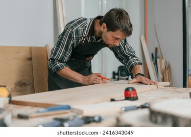 Master of woodcraft male with carpentry tools working in craft workshop. Young carpenter wearing apron making furniture in wood factory workplace, using pencil to mark on timber. Joinery wood concept. - Powered by Shutterstock