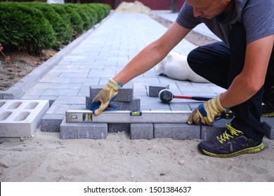 The master in yellow gloves lays paving stones in layers. Garden brick pathway paving by professional paver worker. Laying gray concrete paving slabs in house courtyard on sand foundation base. - Shutterstock ID 1501384637