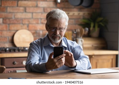 Mastering Modern Gadget. Focused Senior Man Pensioner In Glasses Sit By Table Kitchen Hold Mobile Phone Dial Number Search Data Online. Serious Elderly Grandfather Make Call On Cell Check Email In App