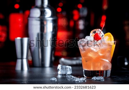 Matador alcoholic cocktail drink with scotch whiskey, white vermouth, liqueur, bitter, grenadine, orange juice, cocktail cherry and ice. Black bar counter background, steel bar tools and bottles