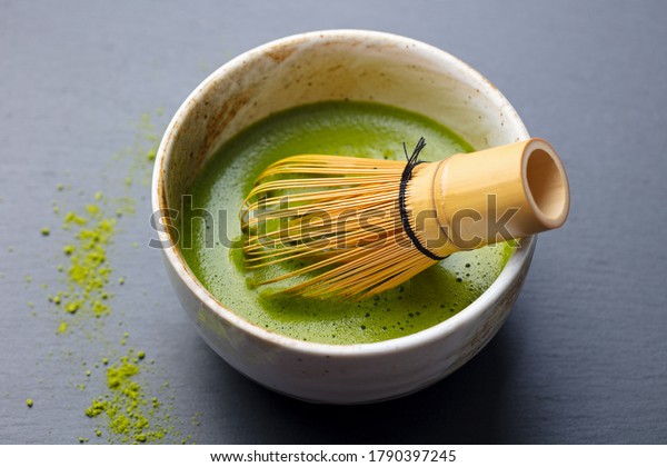 Matcha green tea cooking process in\
a bowl with bamboo whisk. Grey background. Close\
up.