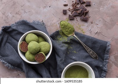 Matcha powder healthy energy balls with dates, nuts and raw ingredients served on a brown textured surface next to chopped chocolate and spoon with matcha powder, shot from the top, healthy dessert