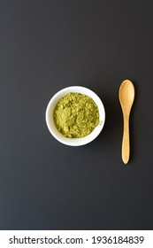Matcha powder in a white bowl and a wood spoon on a black background. Vertical, top view. 
