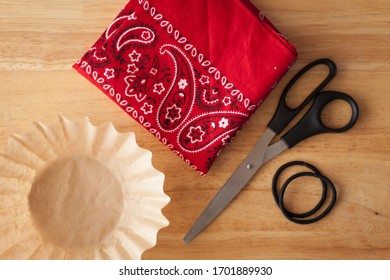 Materials For An Emergency Cloth Face Covering, An Easy DIY Face Mask. Flat Lay Of A Bandanna, Scissors, Coffee Filter, And Hair Ties