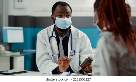 Maternity Specialist And Pregnant Woman With Face Mask At Checkup Visit, Meeting To Talk About Pregnancy. Future Mother With Baby Bump Asking For Medical Advice From Physician At Cabinet Desk.