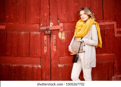 Maternity Woman Holding The Belly With Love And Expecting Baby. Young Pregnant Woman Posing Against The Street Wall. Wearing Fashion Coat, Yellow Scarf Aand Warm Sweater. 