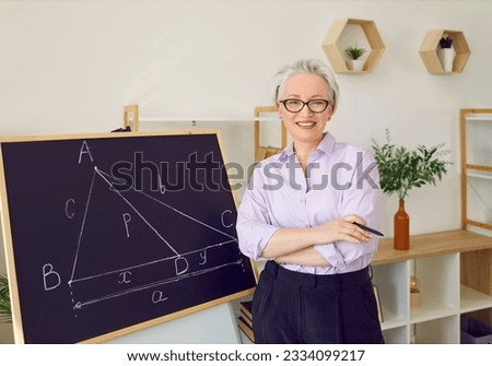 Maths teacher in front of blackboard in school classroom. Happy beautiful elegant middle aged woman in shirt and glasses standing by chalk board with geometry theorem, looking at camera and smiling