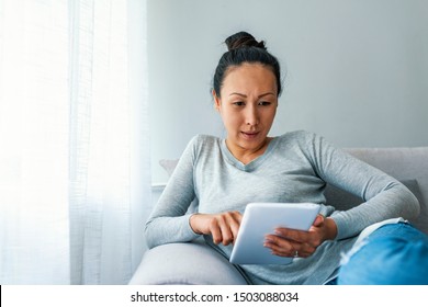 Matue Asian Woman Using Digital Tablet Whilst Relaxing On Sofa. Mature woman using digital tablet at home. Downtime in the digital age. Woman Using Tablet Computer On Sofa - Powered by Shutterstock