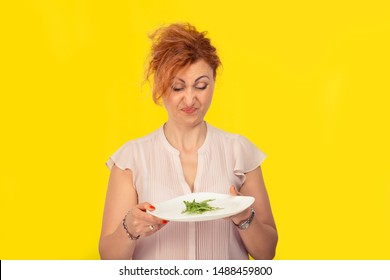 A mature 40 years old woman hates her vegetarian diet concept. Woman looking with disgust to the green salad on her plate isolated over yellow background
