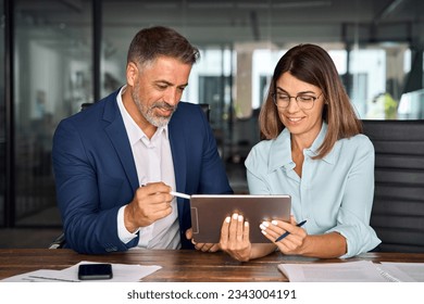 Mature 50s age Latin business man mentoring mid age European business woman discussing project on tablet in office. Two colleagues, group of partners, professional business people working together. - Powered by Shutterstock