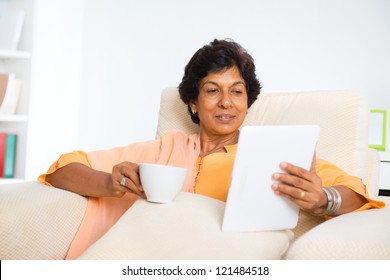 Mature 50s Indian Woman Drinking Coffee / Tea And Using Digital Computer Tablet At Home