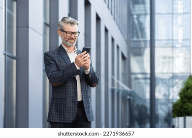 Mature adult businessman outside office building walking and using smartphone, boss typing message in online application on phone, smiling and happy reading news online. - Powered by Shutterstock