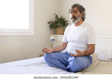 Mature adult man relaxing, doing meditation sitting on bed at home. Male meditating in the morning. Copy space. Wellness and mental health concept. - Powered by Shutterstock