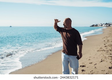 A mature adult man, wearing a T-shirt and jeans, happily walks along the seashore. A smiling man walks along the beach on a sunny day. A bald man with a beard walks along the seashore. - Powered by Shutterstock