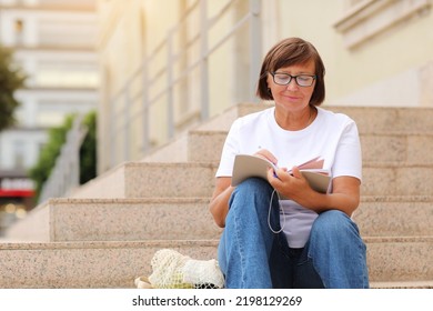 Mature Adult Student Woman In Glasses Writing In Notebook, Sitting On The Stairs At College Outdoors. Happy And Smiling Senior Female In White T Shirt. Distance Studying Online Education And Work