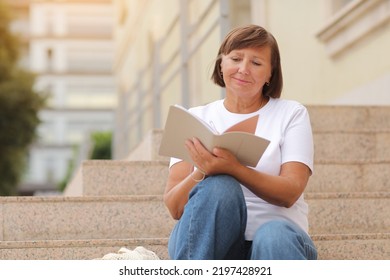 Mature Adult Student Woman Writing In Notebook, Sitting On The Stairs At College Outdoors. Happy And Smiling Senior Female In White T Shirt. Distance Studying Online Education And Work. Copy Space.