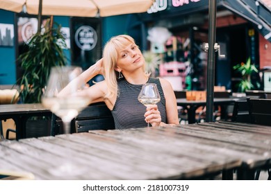 Mature Adult Woman Sitting In Bar Outdoors With Wine Glasses And Blurry Restaurant Background Scene, Drinking White Wine And Eating Cheese. Summer Sunny Day On Patio. People Lifestyle