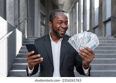 Mature African American businessman is happy about the successful results of financial activity, man is holding in his hands a smartphone and cash American money dollars, outside the office building.