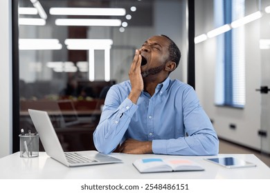 Mature African American businessman yawning at office desk with open laptop. Depicts workplace fatigue, stress, and need for rest. Blue shirt contrasts modern professional environment. - Powered by Shutterstock