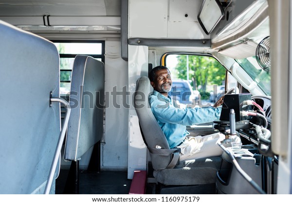 mature african american driver sitting at bus and\
looking at camera