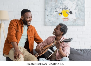 Mature african american father playing acoustic guitar near son on couch at home - Powered by Shutterstock