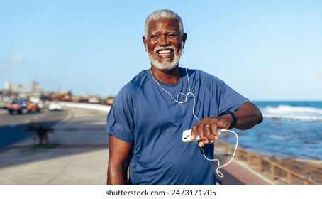 Mature African American man enjoying exercise by the seaside, using fitness watch and listening to music. Sunny day with ocean background. - Powered by Shutterstock