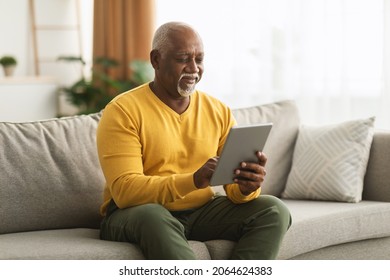 Mature African American Man Using Digital Tablet Sitting On Sofa At Home. Senior Male Browsing Internet On Computer Reading And Scrolling News Feed Online. People And Gadgets - Powered by Shutterstock