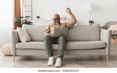 Mature African American Man Using Laptop Pointing Remote Controller Up Turning On Air Conditioner Sitting On Couch In Living Room At Home. Domestic Comfort And Climate Control Concept - Powered by Shutterstock