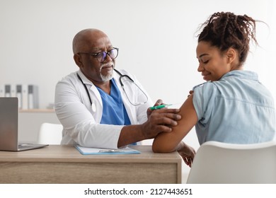 Mature African American Medical Doctor In Glasses Giving Vaccine Shot To Young Female Patient At Clinic. Smiling Black Woman Getting Covid 19 Or Tdap Vaccine, Looking At Syringe And Shouder