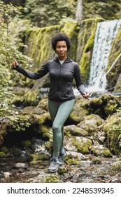 Mature African American woman hiker crossing a stream near the waterfall while exploring in the mountains and enjoying outdoor training. - Shutterstock ID 2248539345
