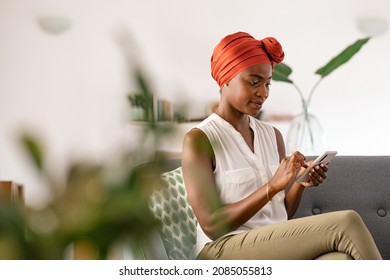 Mature African American Woman Sitting At Home On Couch Using Smart Phone. Beautiful Mid Adult Woman Typing On Mobile Phone. Smiling Ethnic Woman With Traditional Head Wrap Using Cellphone.