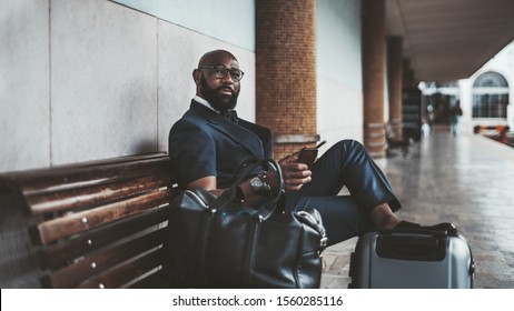 A Mature African Bald Bearded Man Entrepreneur Waiting For A Train To Start His Business Trip And Sitting On The Wooden Bench At The Platform Of A Railway Station With A Smartphone And Bags Near Him