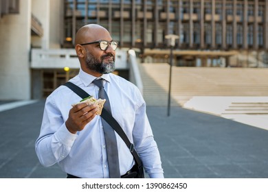 Mature African Businessman Eating Sandwich Outdoor. Senior Entrepreneur Standing Outside Office Building, Looking Away While Enjoying Vegetable Sandwich. Business Man Having Lunch On The Move.