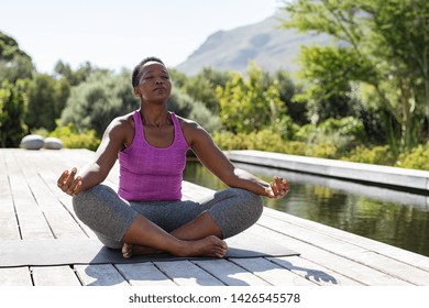 Mature african woman practicing yoga and meditates near swimming pool outdoor. Fitness black lady sitting in lotus pose with closed eyes. African american woman meditating at poolside with copy space.