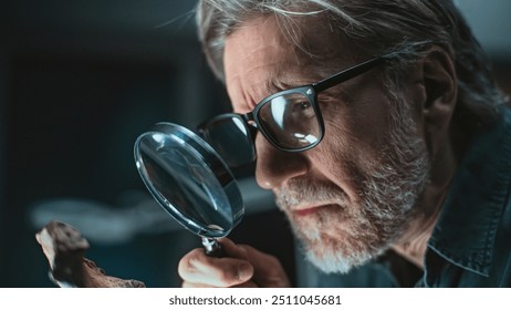 Mature archaeologist examines ancient extinct human or animal remains using magnifying glass. Scientist works with fossil, does scientific bone examination and research in archaeological lab at night. - Powered by Shutterstock