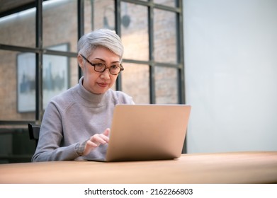 Mature Asian Business Woman Wears Glasses Using Laptop Computer Sit At Workplace Desk. Happy Senior Older Employee 50s Businesswoman
