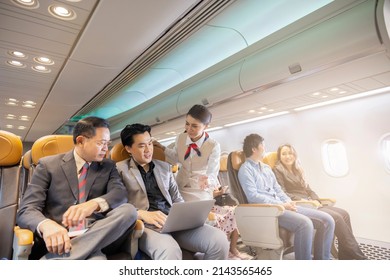 Mature Asian Businessman And Young Businessman Having Discussion, Working Together On Laptop Sitting In Cabin Flight Travel In Economic Class While Flight Attendants Provide Services To Passengers.