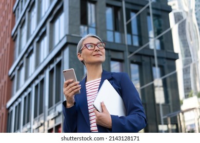 Mature asian businesswoman wearing eyeglasses using mobile phone holding laptop standing on urban street. Portrait of successful manager waiting for taxi looking away outdoors