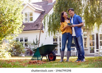 Mature Asian Couple Taking A Break With Hot Drinks Whilst Tidying Garden With Rake And Barrow