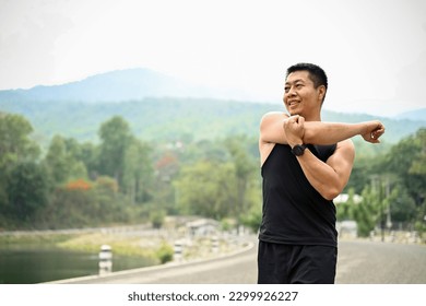 A mature Asian man in sportswear warming up his body before workout, stretching his arms muscle, exercising at a park. Lifestyle concept